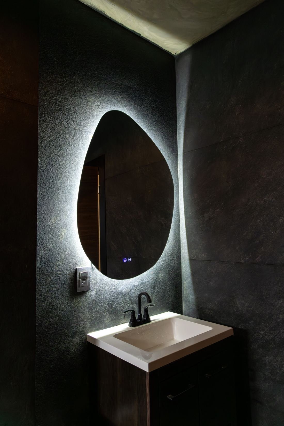 Baño con muros de piedra negra y espejo orgánico retroiluminado